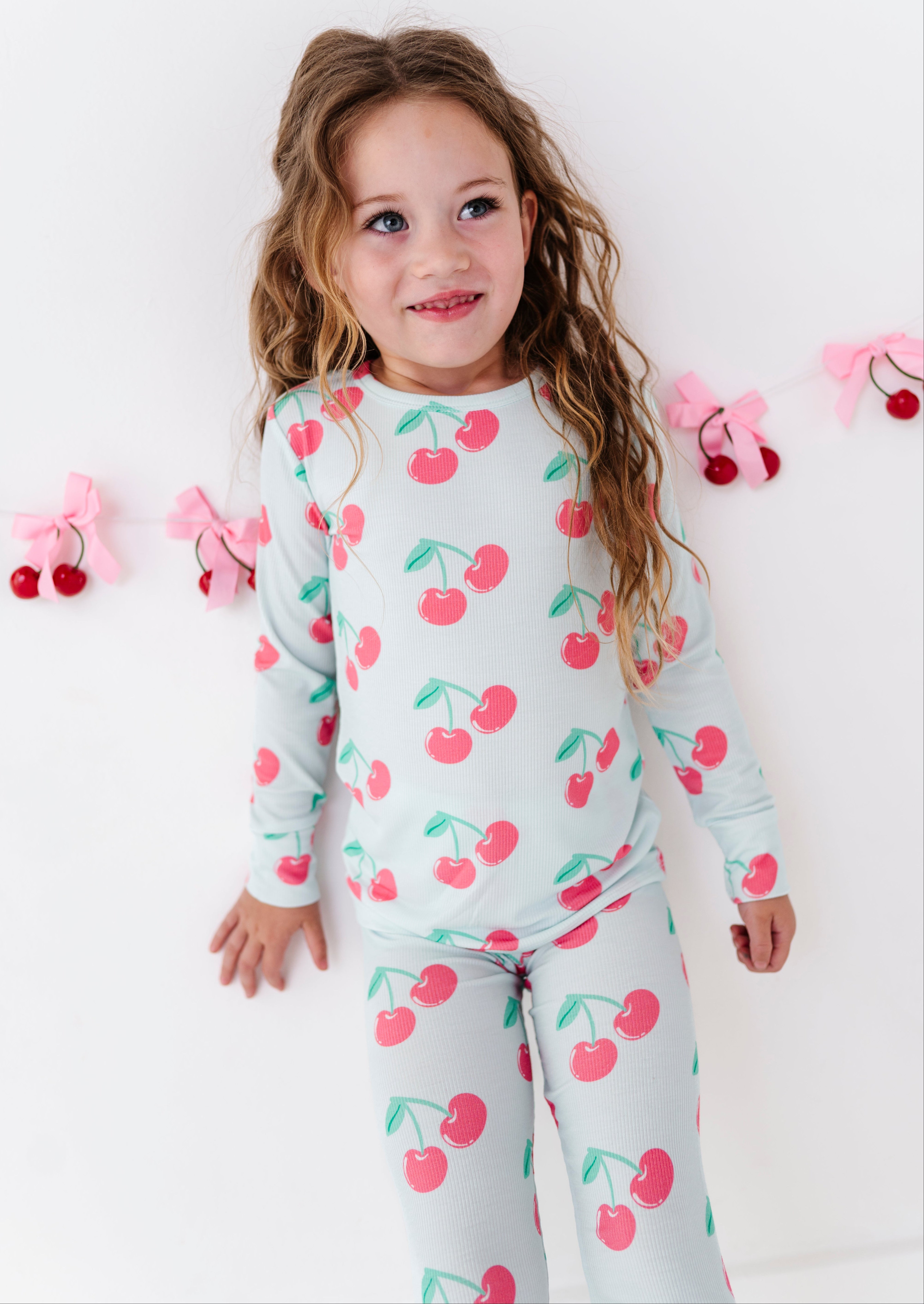 A young girl with long wavy light brown hair is wearing a light gray long-sleeve pajama set with a pink and green cherry print, standing against a white wall with pink bow and cherry decorations.